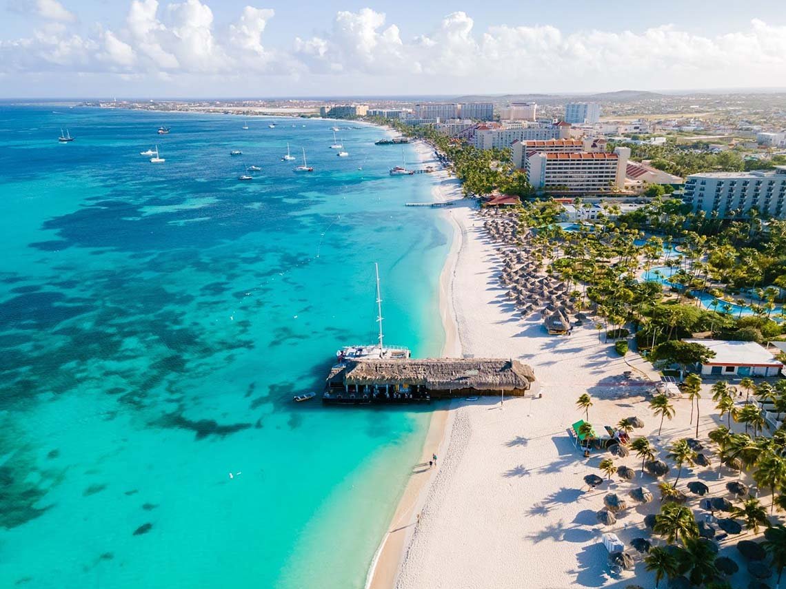 An aerial view of Aruba's long sandy beach with palm trees and huts.  