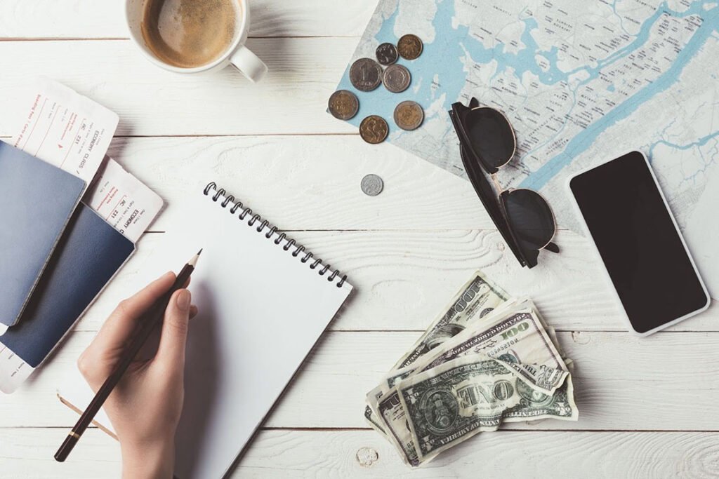 A cropped shot of a woman planning a trip to Aruba with a notebook, map, money, passports, sunglasses, coffee, and a smartphone.