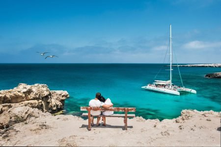 A couple sitting on a bench by the beach watches a catamaran sail on Aruba's waters.